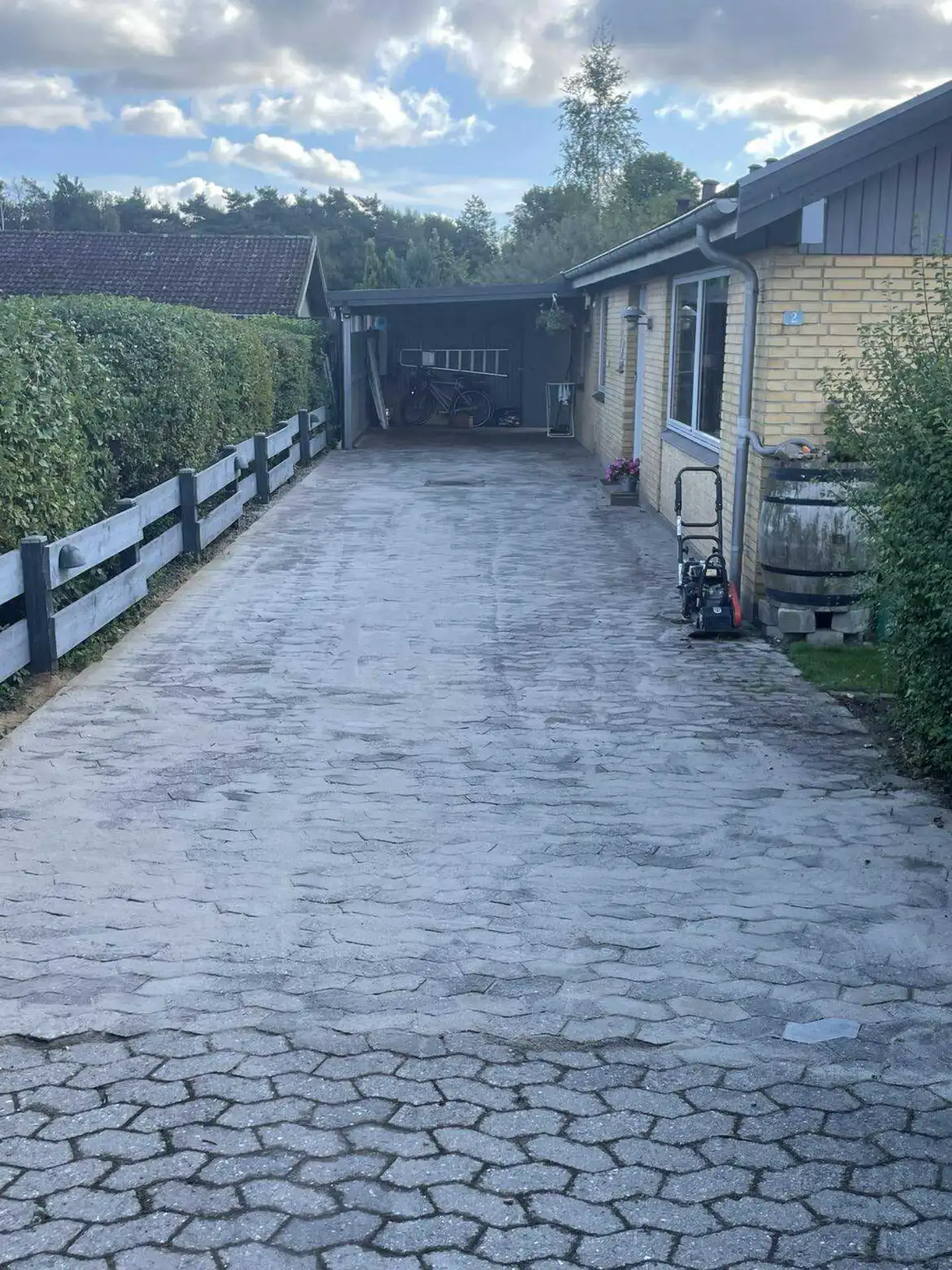 Højt belagt indkørsel med brosten, sidebygning og have i baggrunden, under en skyet himmel.